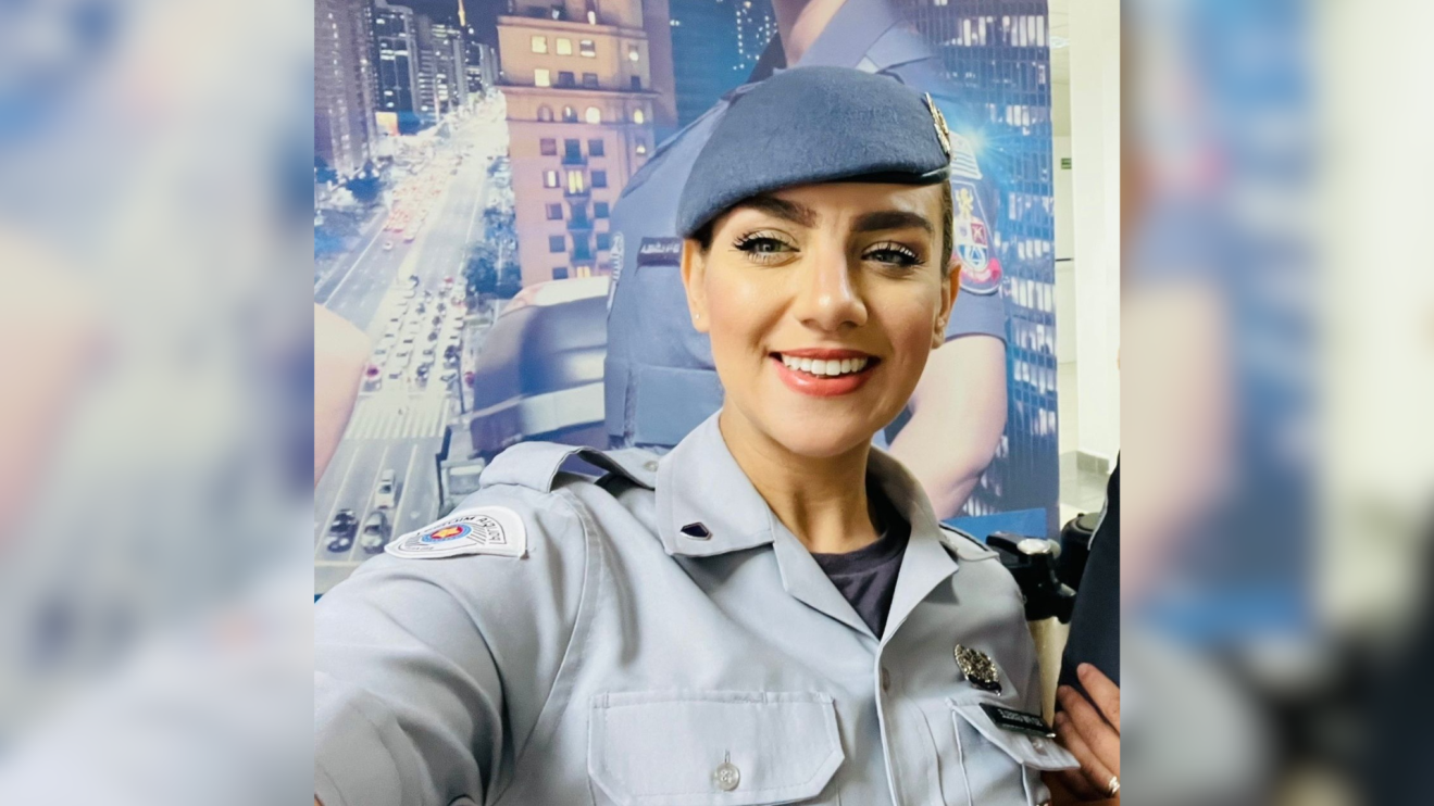 A imagem mostra uma mulher sorrindo, vestindo uniforme policial e tirando uma selfie em ambiente urbano iluminado ao fundo. No contexto da reportagem, o retrato humaniza a vítima de feminicídio e reforça o impacto real das narrativas misóginas que naturalizam relações de poder e violência contra mulheres. Gisele Alves Santana